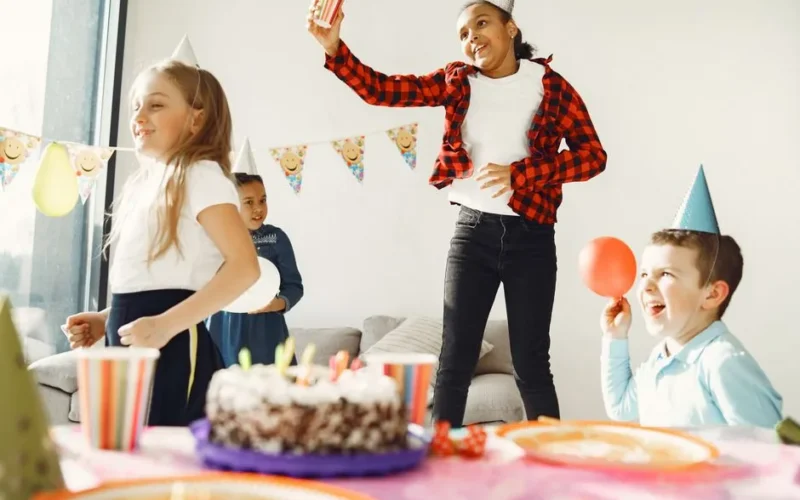 Children having fun at a birthday party with hats, cake, and balloons indoors.