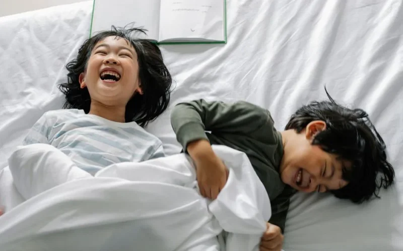 From above of laughing Asian boy and girl lying on bed with book under white blanket in morning in bedroom
