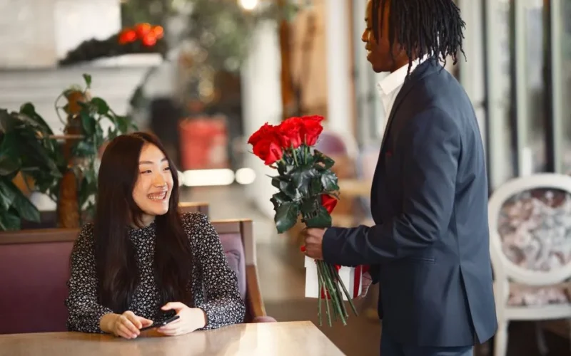 A man in a black suit presents red roses to a smiling woman at a cozy cafe.