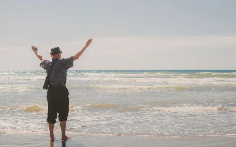 A man joyfully raising arms at the beach, soaking up a sunny, carefree day by the ocean.