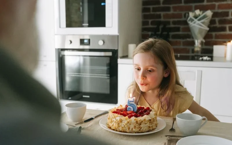 A young girl blows out a birthday candle on a cake during a fifth birthday celebration indoors.