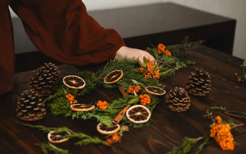 A person arranges a natural Christmas wreath with pinecones and dried orange slices on a wooden table.