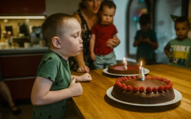 Young boy celebrating birthday by blowing candles on chocolate cake with raspberries.
