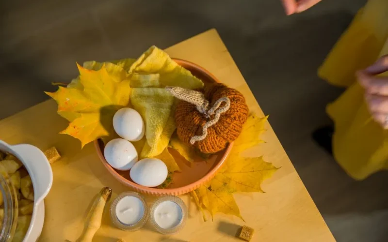 Warm autumn setting featuring a knitted pumpkin, eggs, and candles on a rustic table with fall leaves.