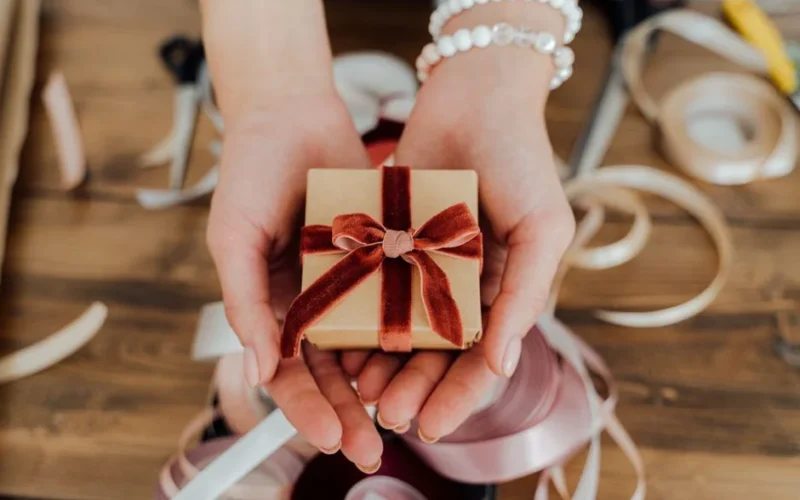 Hands presenting a small gift box wrapped with red velvet ribbon, surrounded by crafting materials.