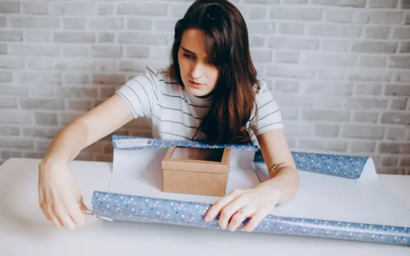 Young woman wrapping a gift box with decorative paper on a table indoors.
