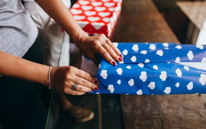 Close-up of a woman wrapping a gift with blue cloud-patterned paper indoors.