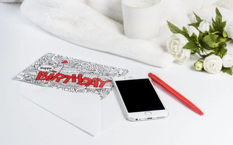 Elegant composition featuring a birthday card, smartphone, candle, and white roses for a stylish greeting.