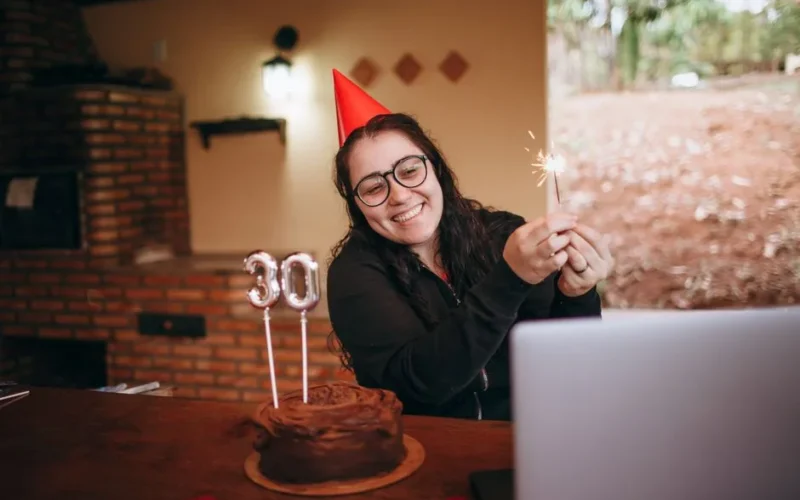 Cheerful woman in party hat holding sparkler with chocolate cake for 30th birthday celebration indoors.