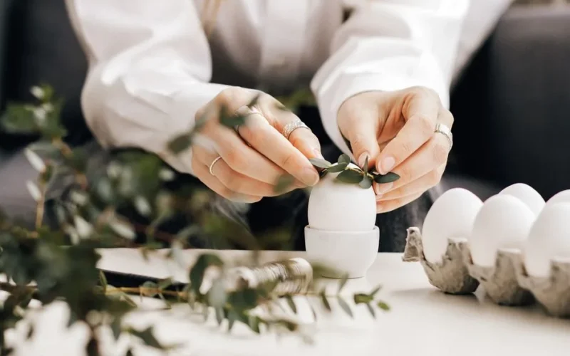 Hands arranging leaves on white eggs for an Easter decoration indoors, creating a natural festive look.