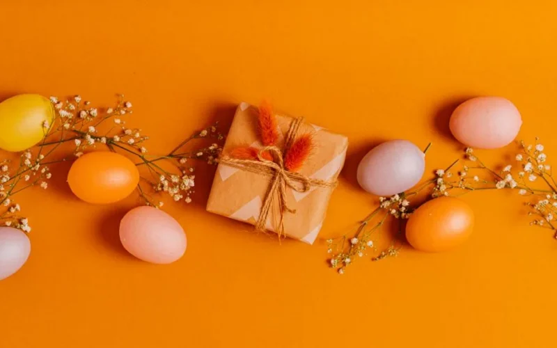 A vibrant arrangement of colorful Easter eggs, flowers, and a wrapped gift on an orange background.
