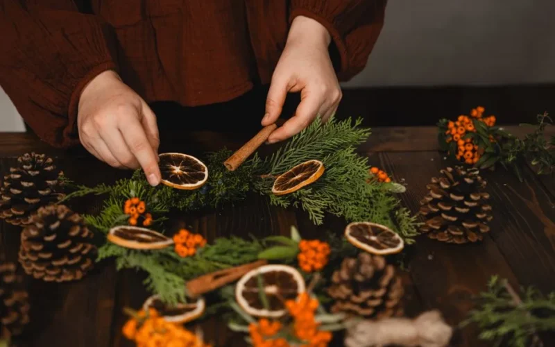 Hands decorating a Christmas wreath with dried oranges and pinecones.