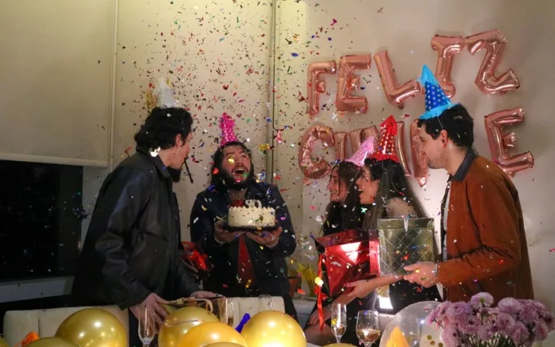 Group of adults celebrating a birthday with cake, gifts, and confetti indoors.