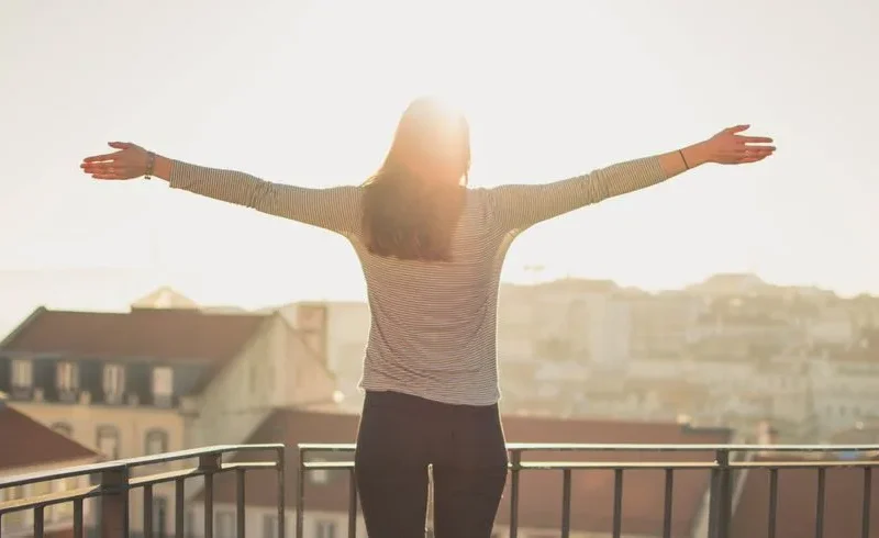 A woman stands with outstretched arms on a sunny balcony, embracing the morning light.