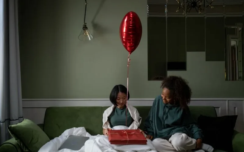 Two friends celebrate with a surprise gift and balloon on a cozy couch setting, creating a warm and joyful atmosphere.