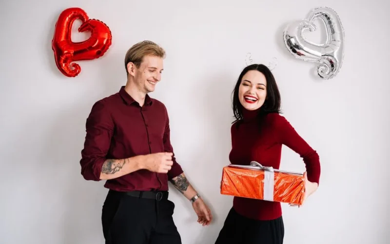 A joyful couple exchanging gifts surrounded by heart-shaped balloons, embracing a romantic celebration.