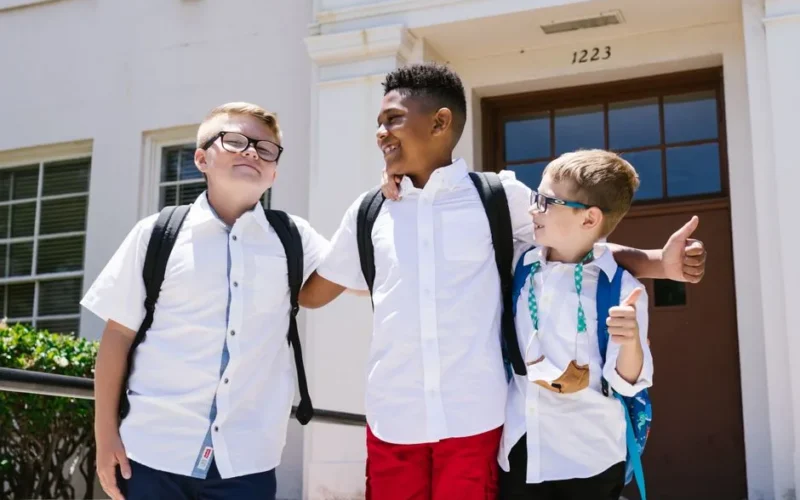 Three smiling schoolchildren with backpacks outside a school building, enjoying their first day back to school.