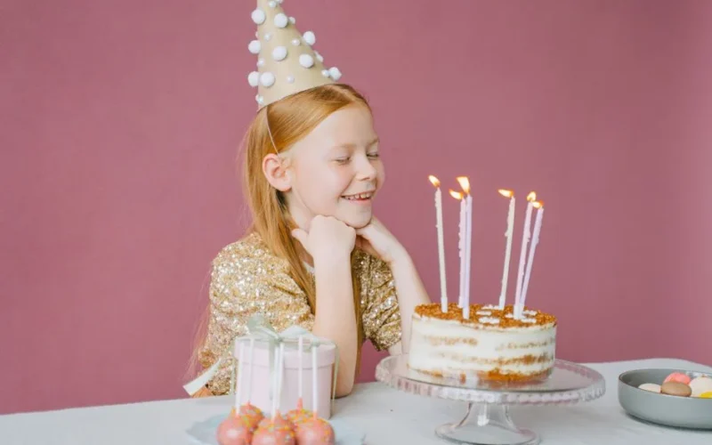 A young girl in a party hat smiles at her birthday cake with candles lit, ready to make a wish.