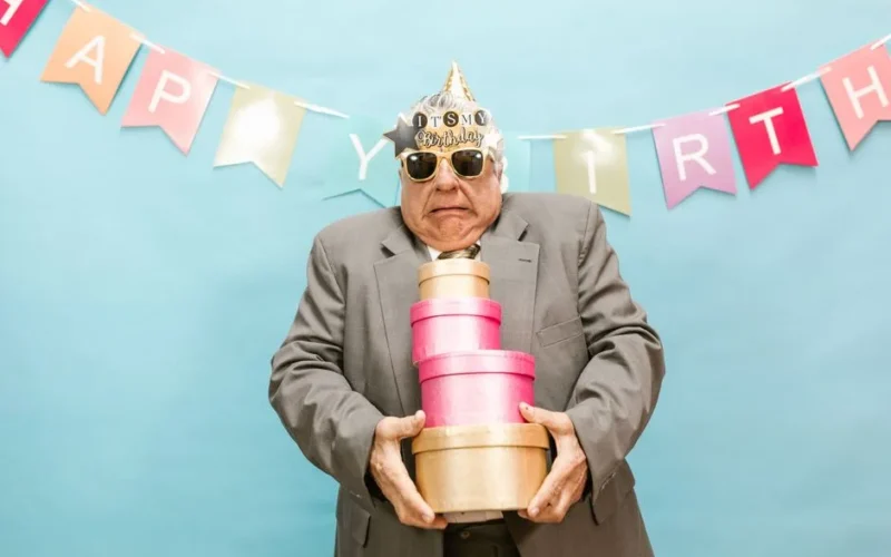 Senior man in party hat holding gift boxes against a colorful birthday banner.