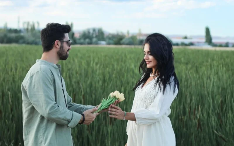 A man and woman share a bouquet, expressing love in a lush green field.