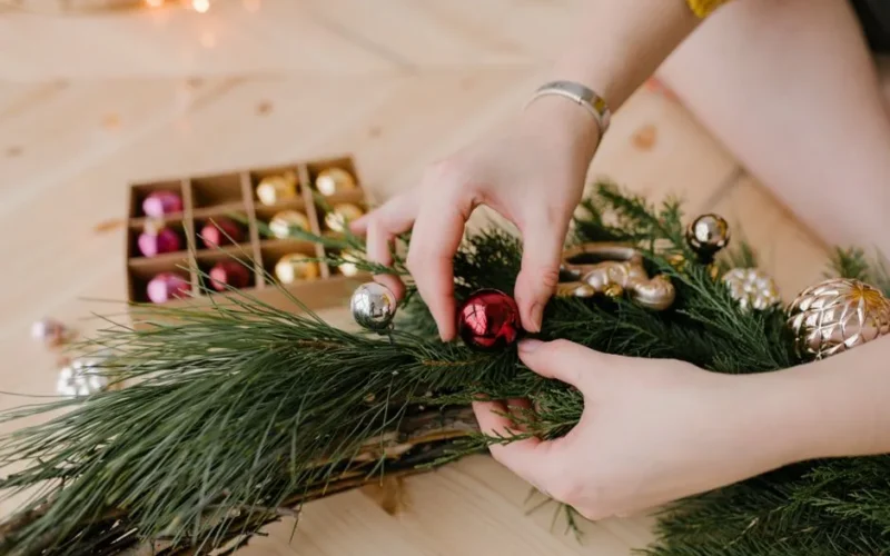 Hands crafting a Christmas wreath with colorful ornaments on a wooden surface.
