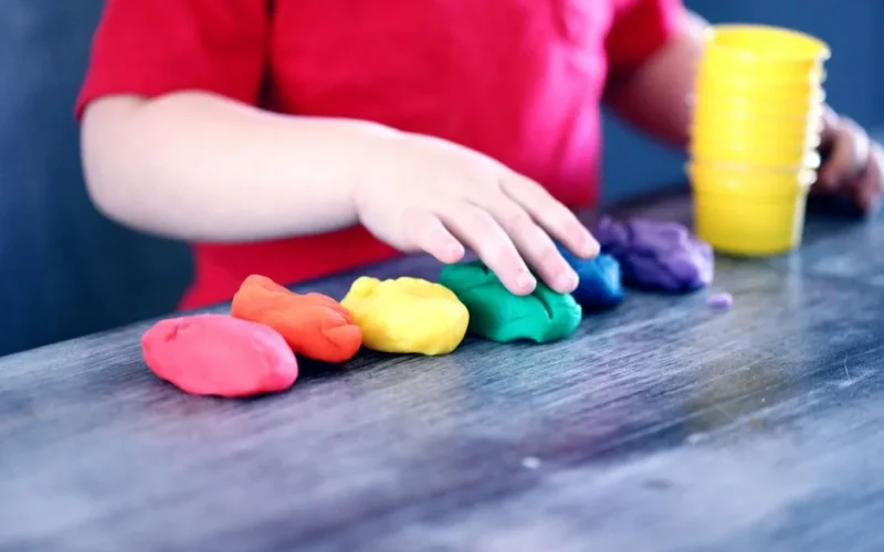 A young child with rainbow-colored modeling clay and stacking cups on a table indoors.