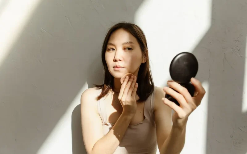 A young woman applying makeup, illuminated by natural light streaming through a window.
