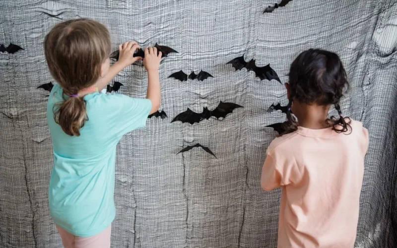 Two kids decorating a wall with bat cutouts, preparing for Halloween indoors.