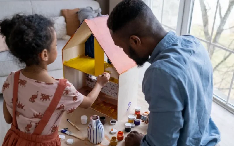 Back view of African American father and little daughter painting with gouache colors wooden toy house for dolls
