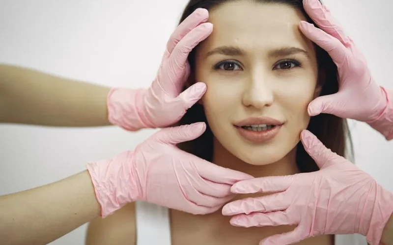 A woman receiving a facial treatment, hands in gloves gently touching her face indoors.