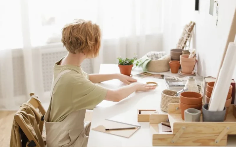 A woman sits in a cozy workspace crafting with flower pots and supplies.