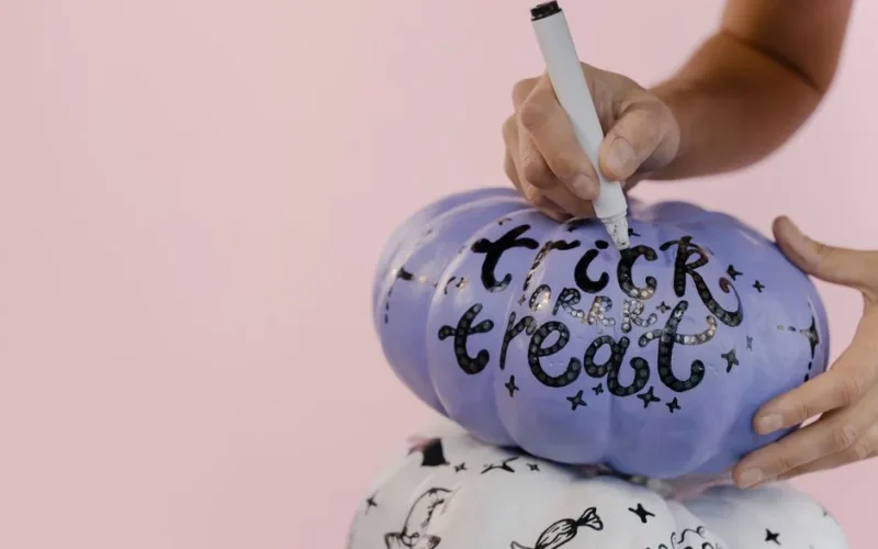 Close-up of hands decorating a blue Halloween pumpkin with "trick or treat" on a pink background.