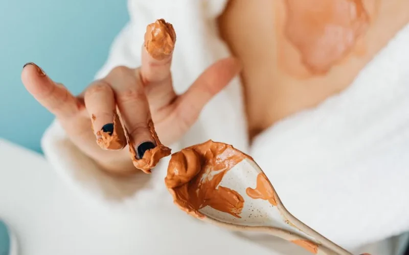 Woman applying homemade mud mask with spoon and fingers in a bathrobe.