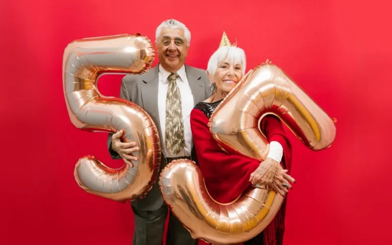 Happy senior couple in formal attire celebrating with golden balloons against a bright red backdrop.