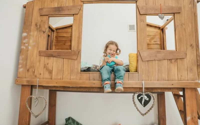 A happy girl sits in a unique wooden bunk bed, smiling and playful.