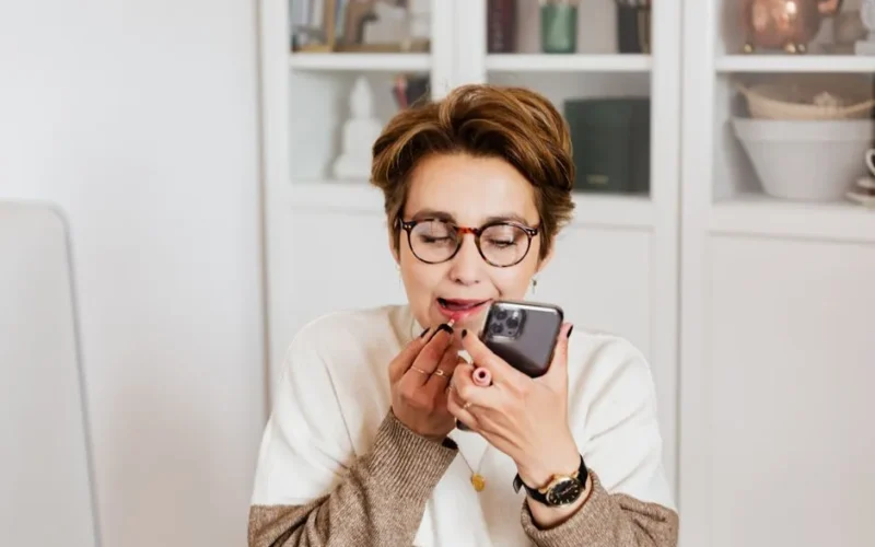 Content stylish female in eyeglasses looking at modern mobile phone front camera and applying lipstick while sitting in cozy light apartment