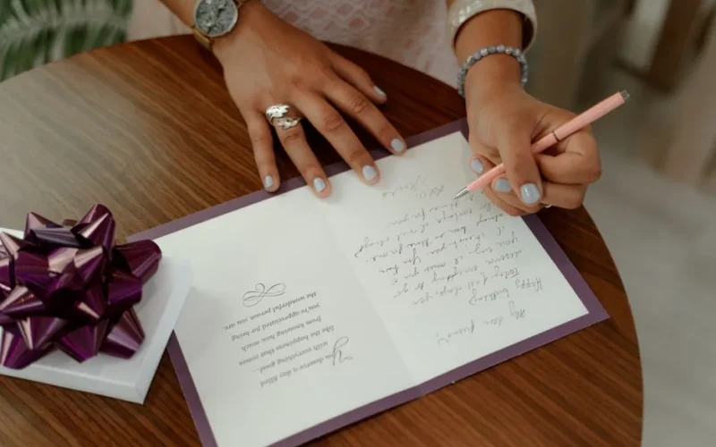 A woman writes a message inside a greeting card on a wooden table with a gift beside.