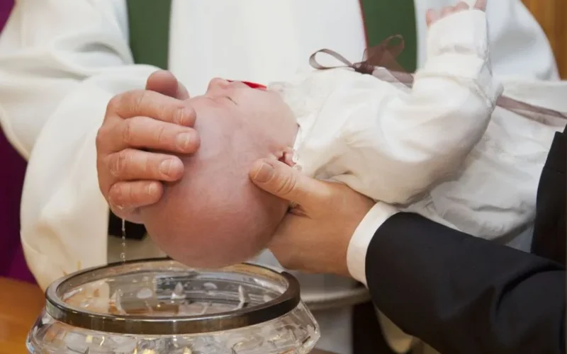 A baby being baptized during a ceremonial Christian baptism, featuring a priest and holy water.