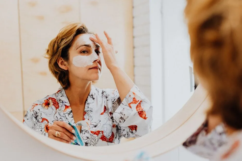 A woman in a towel performing her skincare routine in front of a bathroom mirror.