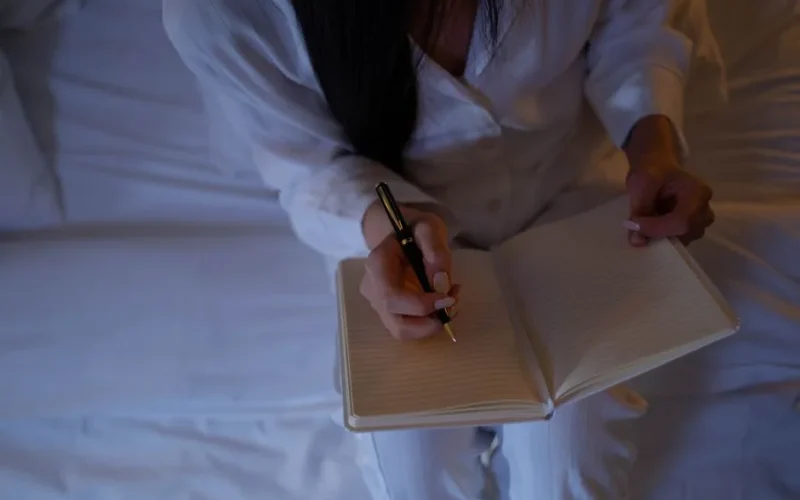 A person is writing in a notebook while sitting on a bed in a dimly lit room.