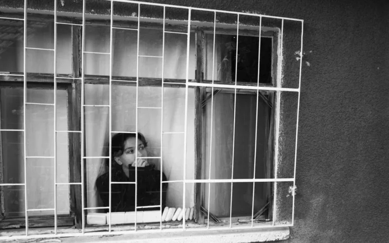 A reflective woman looks out of a barred window, captured in moody black and white tones.