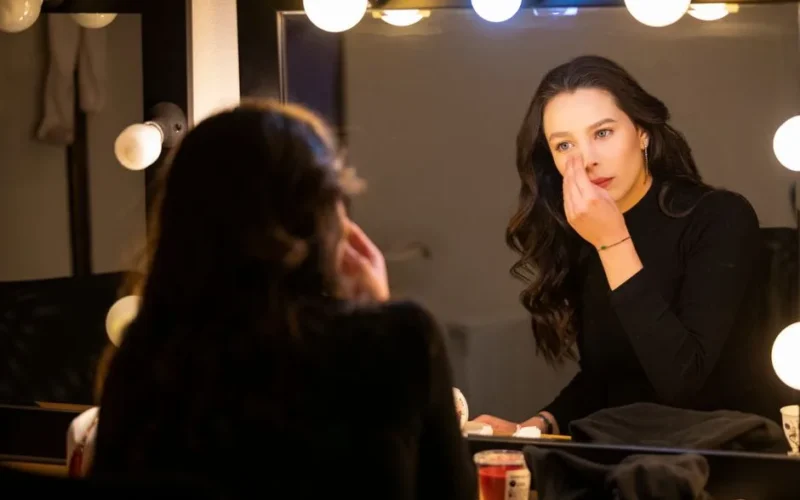Young woman applies makeup in a brightly lit dressing room mirror, creating a focused and elegant atmosphere.