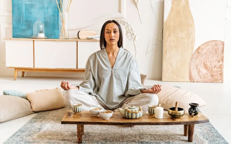 Woman peacefully meditating indoors surrounded by cushions and tea set, promoting tranquility.