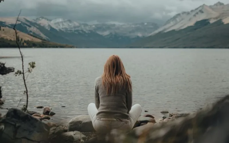 A person sits by a mountain lake, finding peace and solitude in nature's embrace.