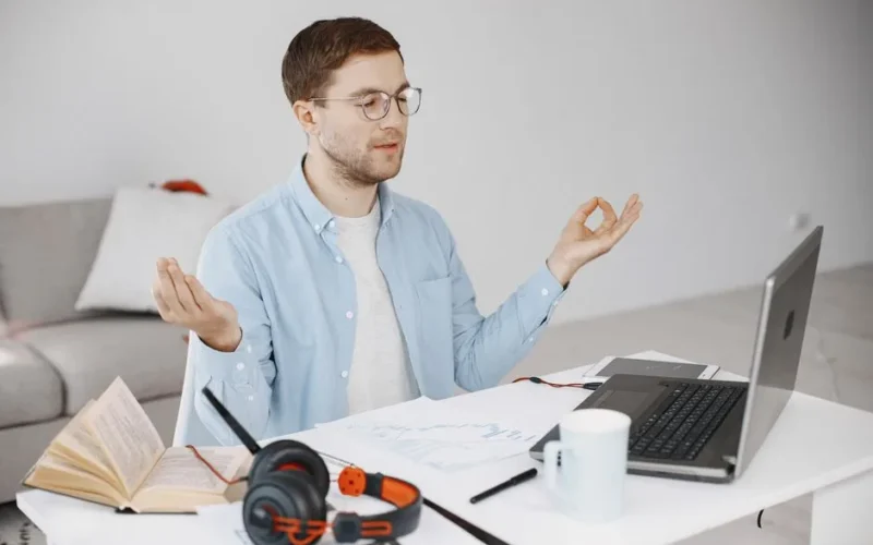 Man practicing meditation at a tidy desk with a laptop, books, and headphones in a home office.
