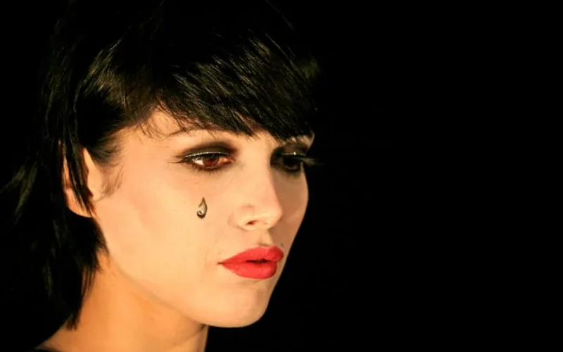 Close-up portrait of a woman with dramatic makeup and a teardrop on her cheek against a black background.