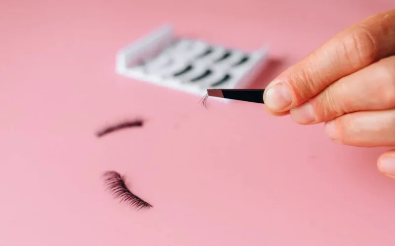 Close-up of applying false eyelashes with tweezers over a pink background.