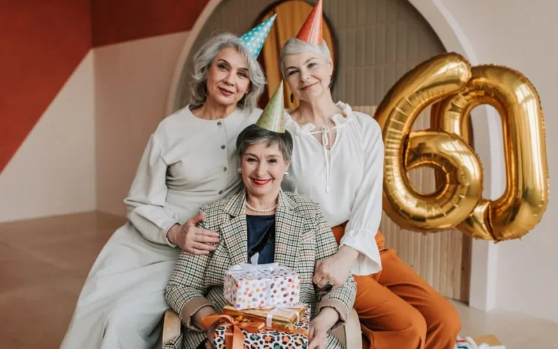 Three elegant women celebrate a 60th birthday with balloons and gifts indoors.