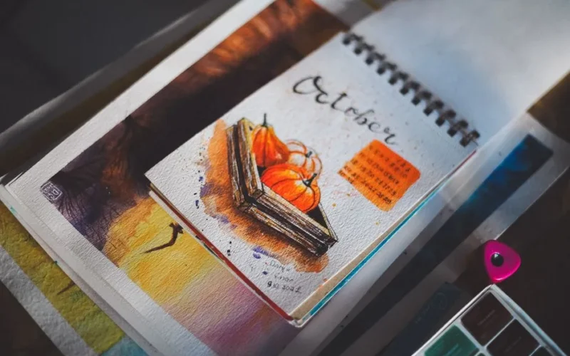 Open sketchbook showing a vibrant October watercolor painting with pumpkins and a calendar.