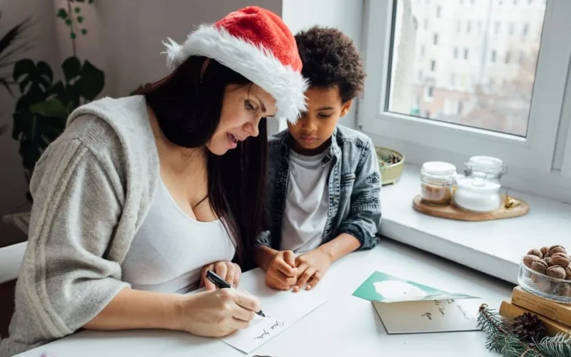 A mother and her son enjoy a festive moment writing Christmas cards by a window.
